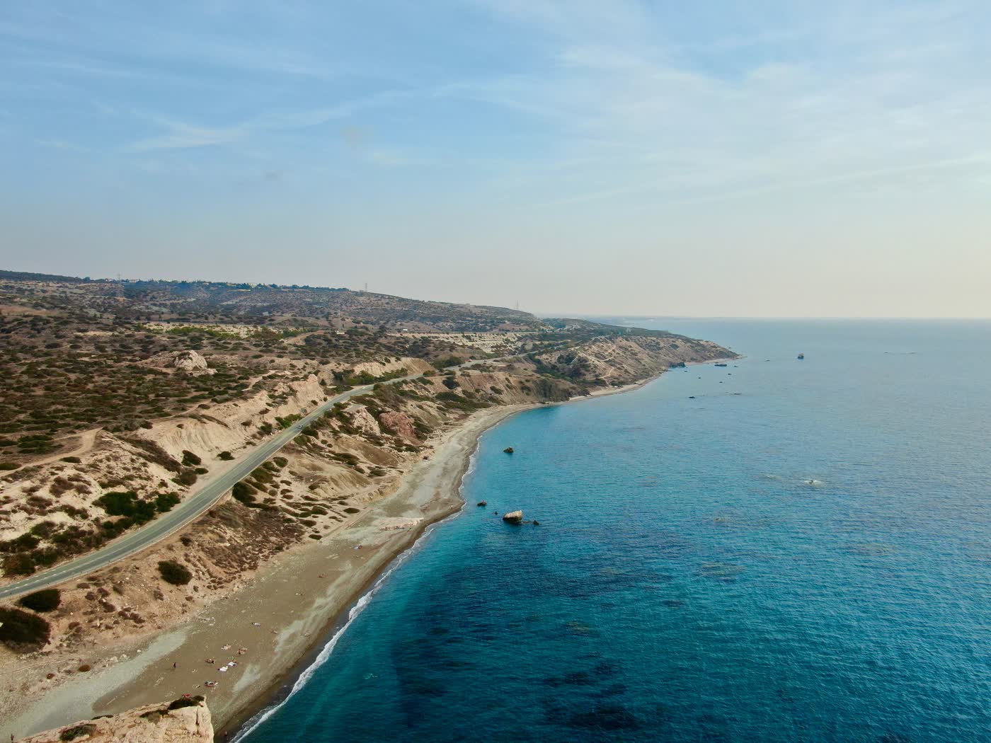 A coast road running along the cliff above a long Cyprus beach, deep blue sea filling the frame