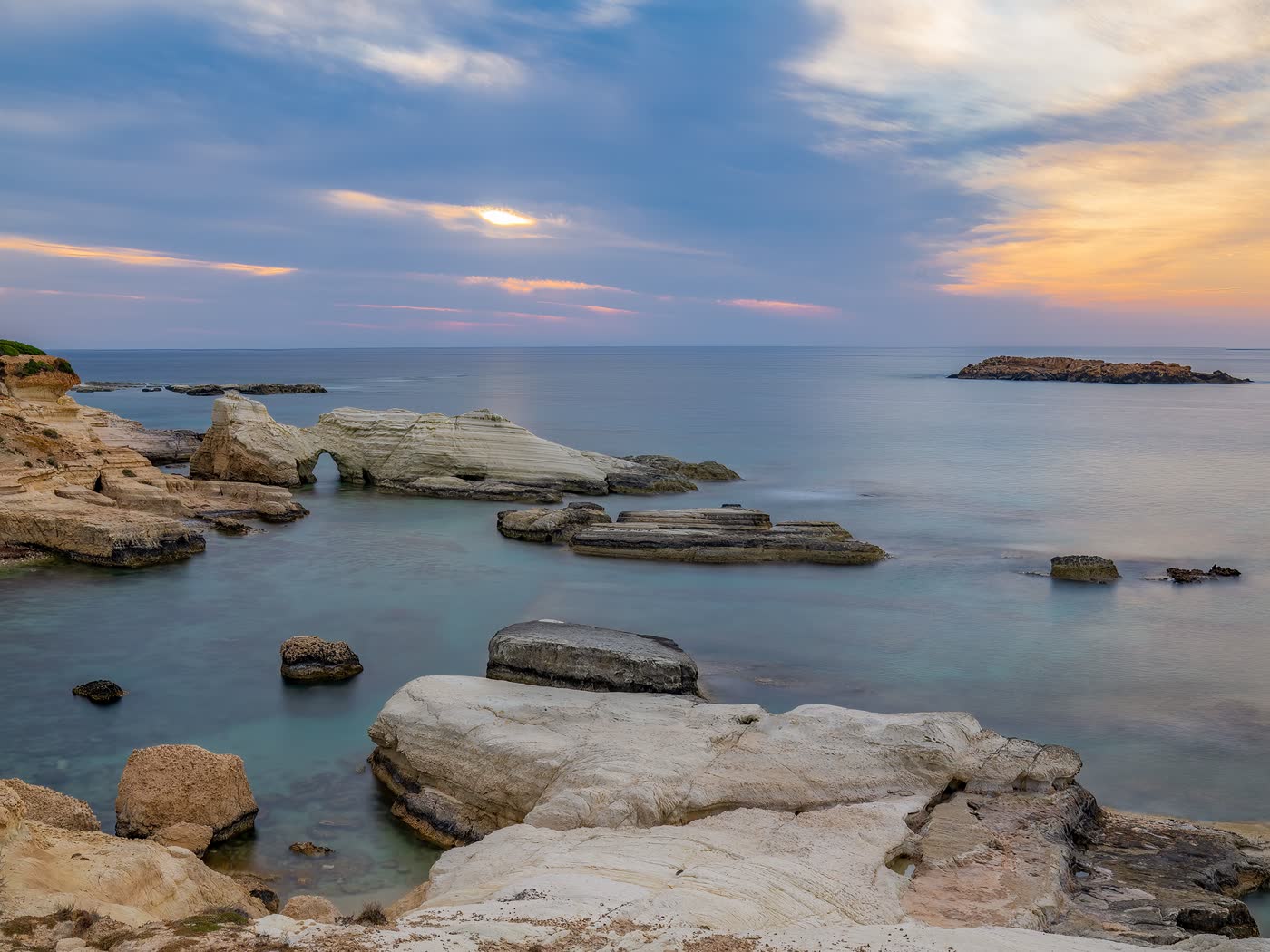 Flat pale rock shelves running into still water on the Cyprus coast under a soft pink and gold dusk