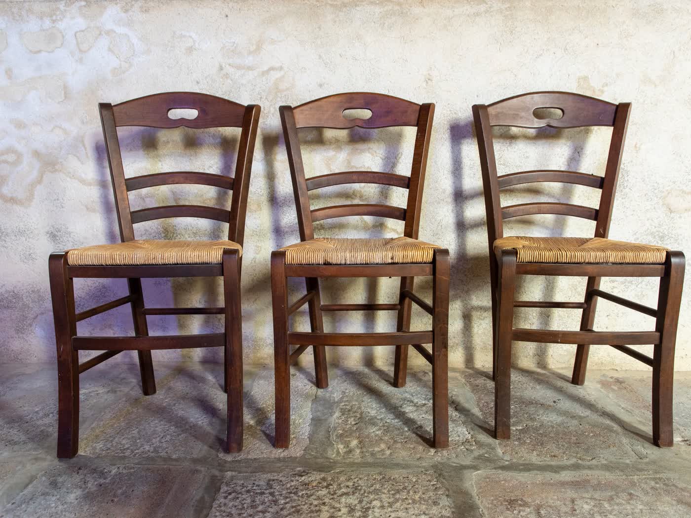 Three rush-seat wooden chairs standing in a row against a bare plastered wall in afternoon sun
