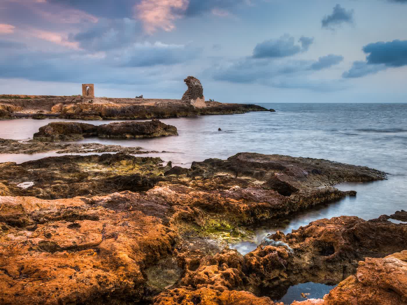 Warm rust-coloured rock shelves on the Cyprus shore at dusk, with a ruined stone arch on the headland