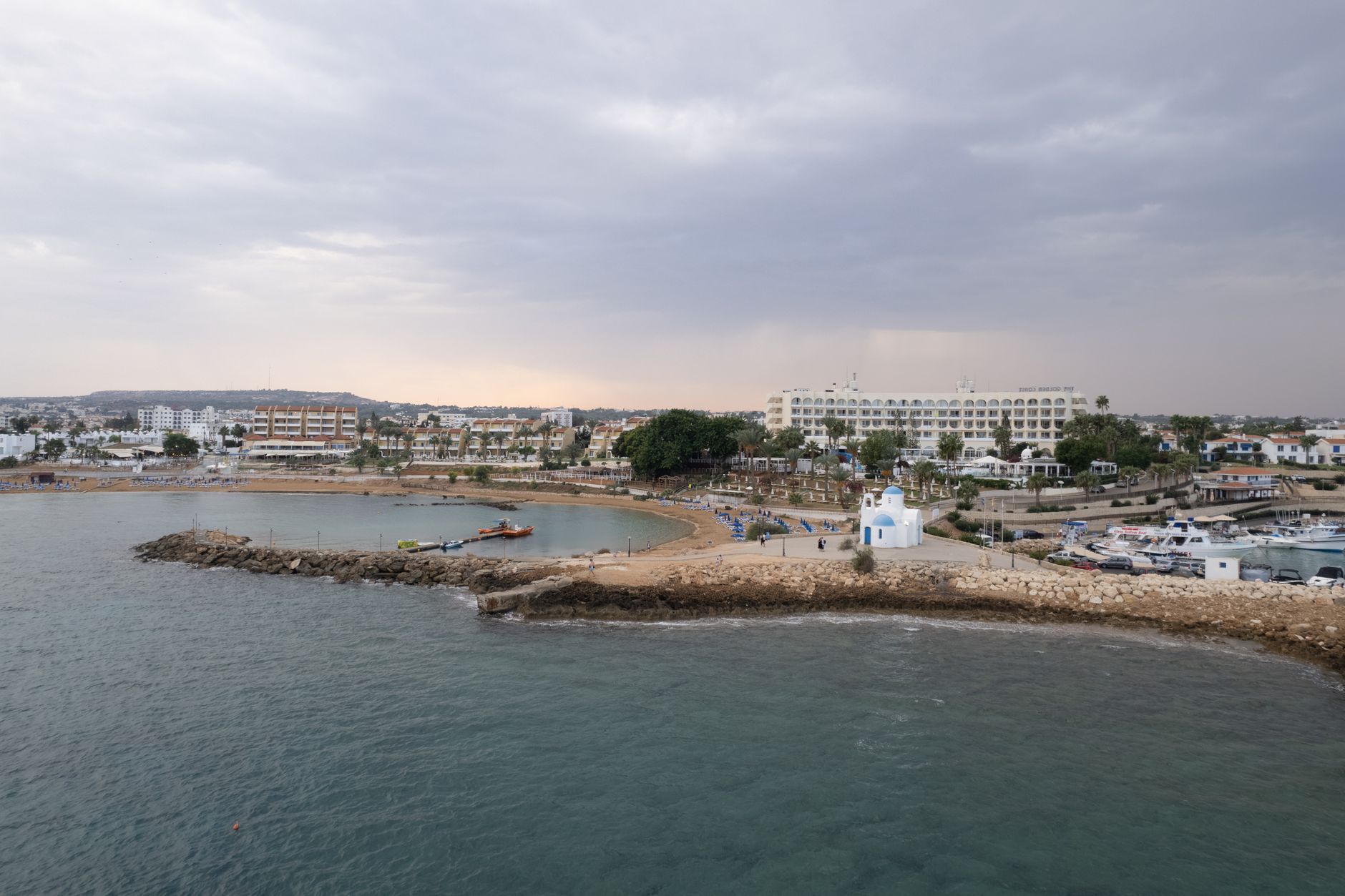 A small blue-domed whitewashed chapel on a rocky Cyprus headland, with a sheltered swimming bay on one side and a fishing harbour on the other