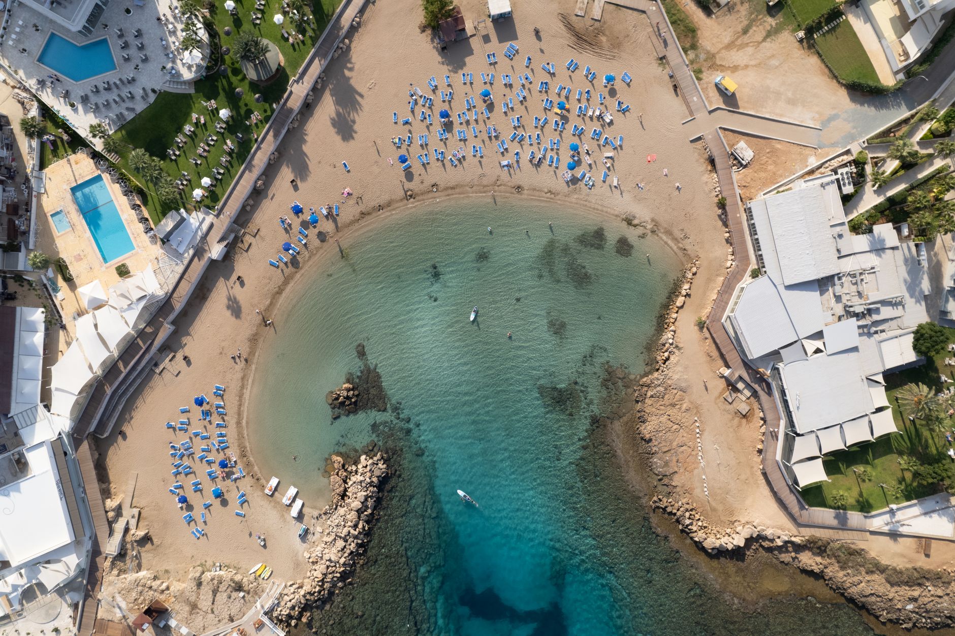 A small sandy cove seen straight down from the air, ringed by hotel terraces with swimming pools, blue parasols and loungers set out on the sand, and two paddleboarders on clear shallow water