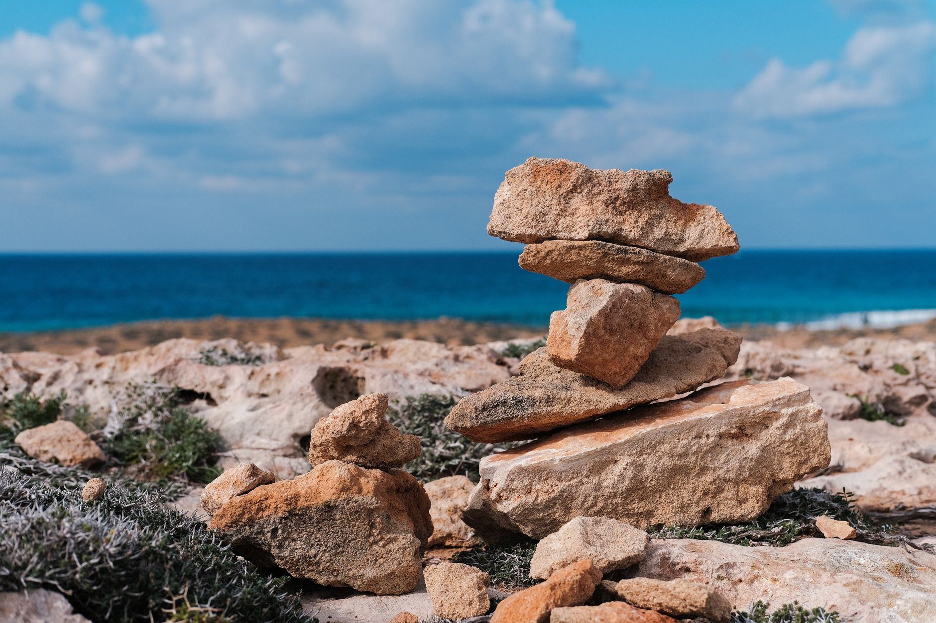 A stack of flat limestone slabs balanced on a rocky coastal ledge in Cyprus, with low scrub, pale sand and deep blue sea behind