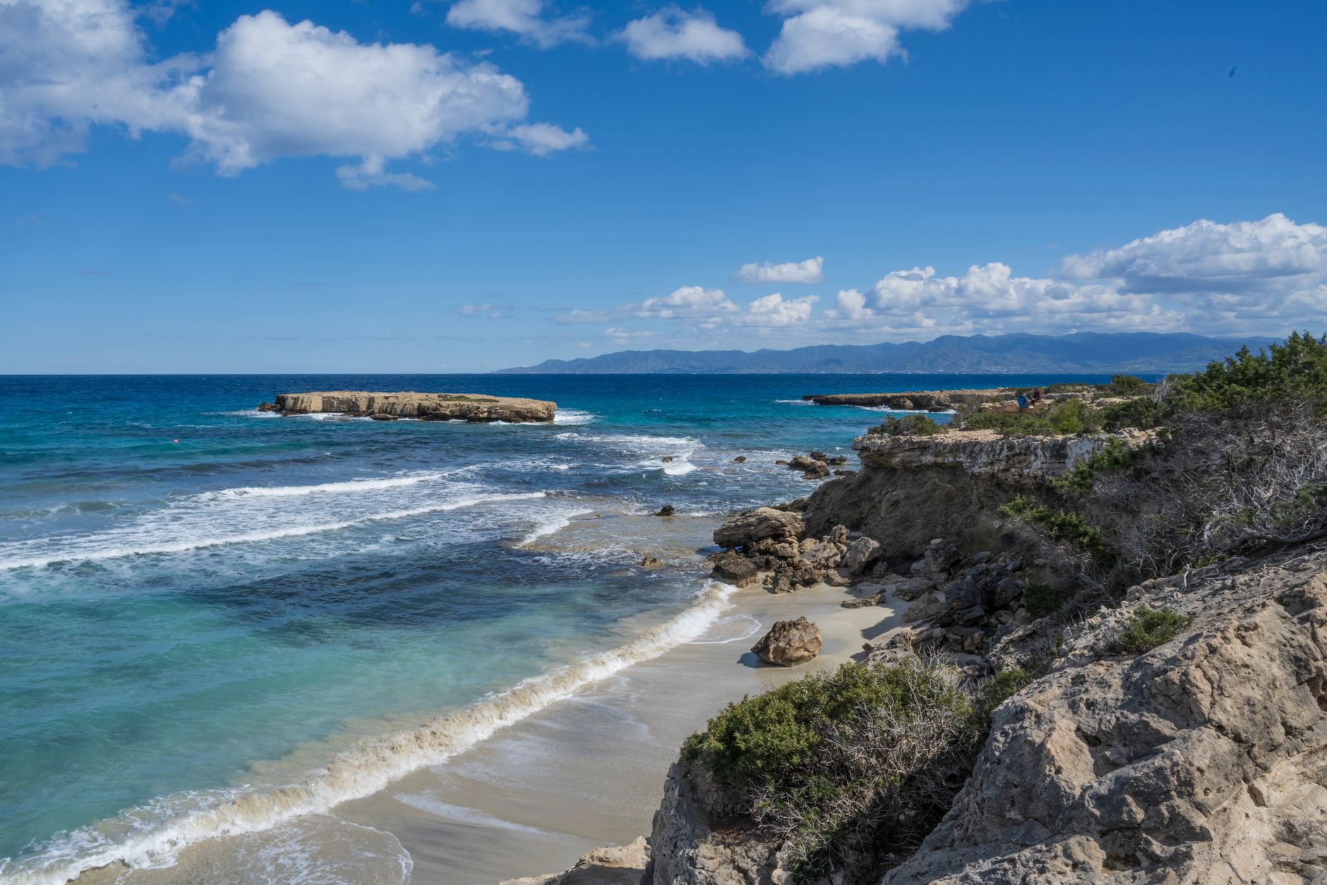A small sandy cove below a low limestone cliff on the Cyprus coast, with turquoise water breaking in white lines and a distant mountain ridge across the bay