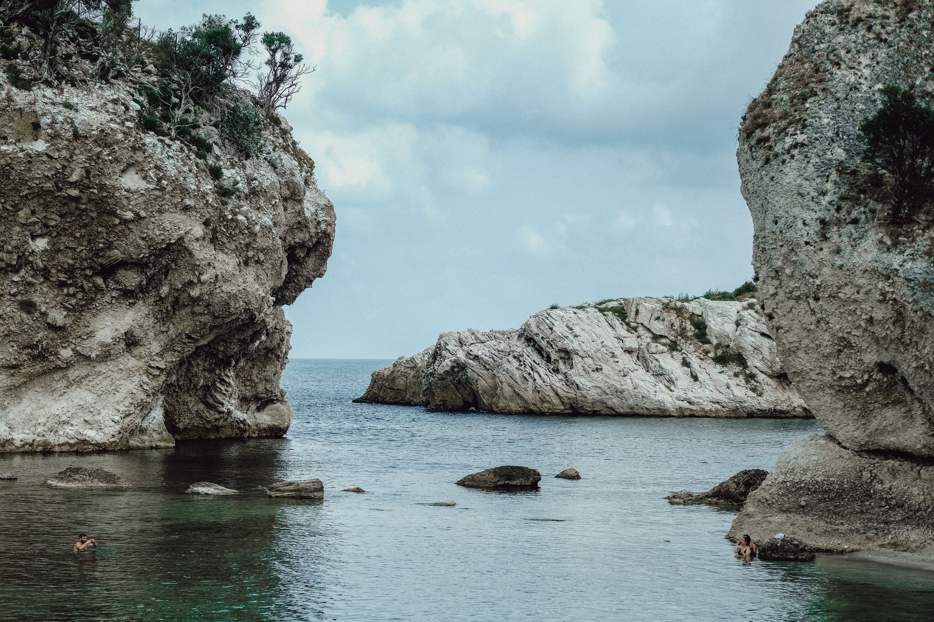 Two swimmers in a sheltered rocky cove in Cyprus, pale limestone headlands rising on both sides of clear green water