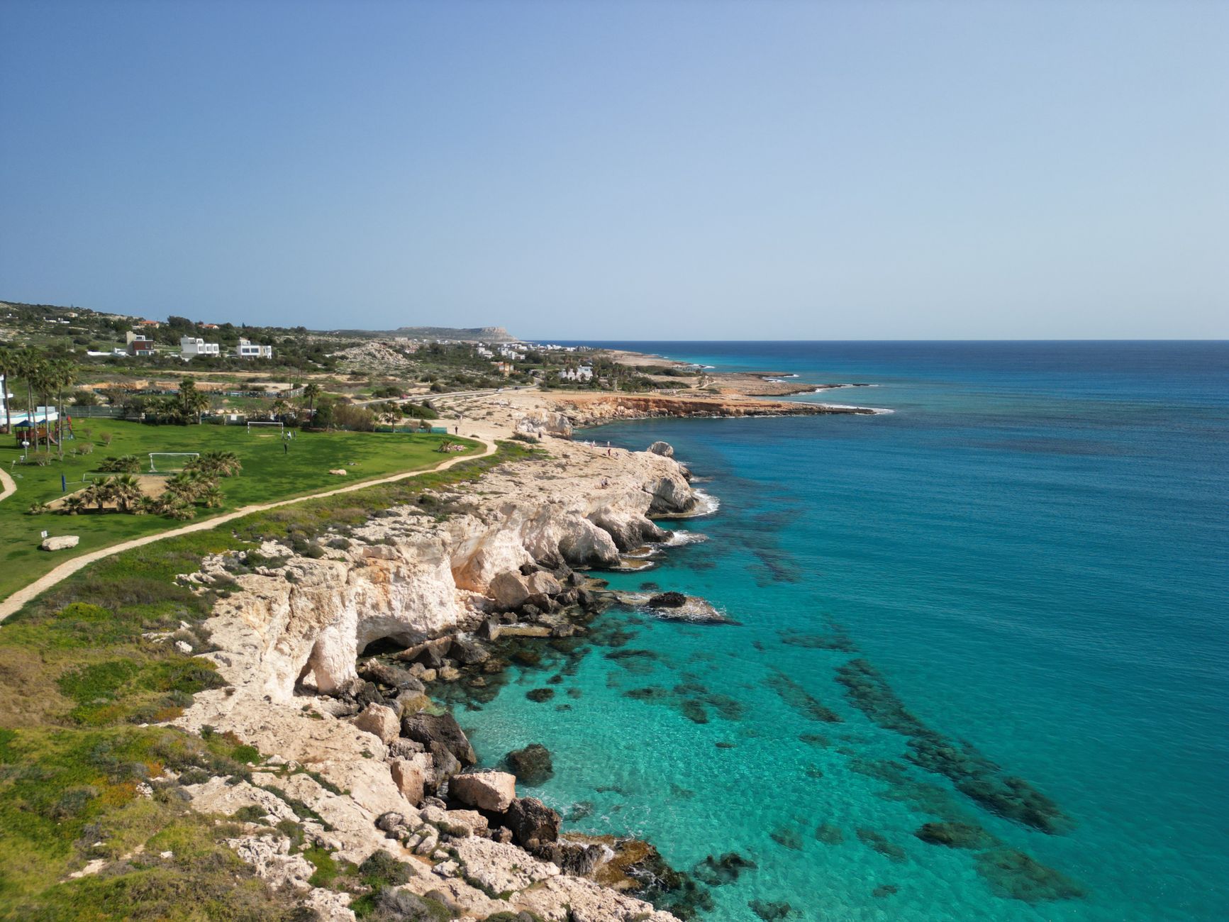 A limestone headland on the eastern Cyprus coast dropping into clear turquoise water, with a green park, palms and low white houses along the shoreline behind it