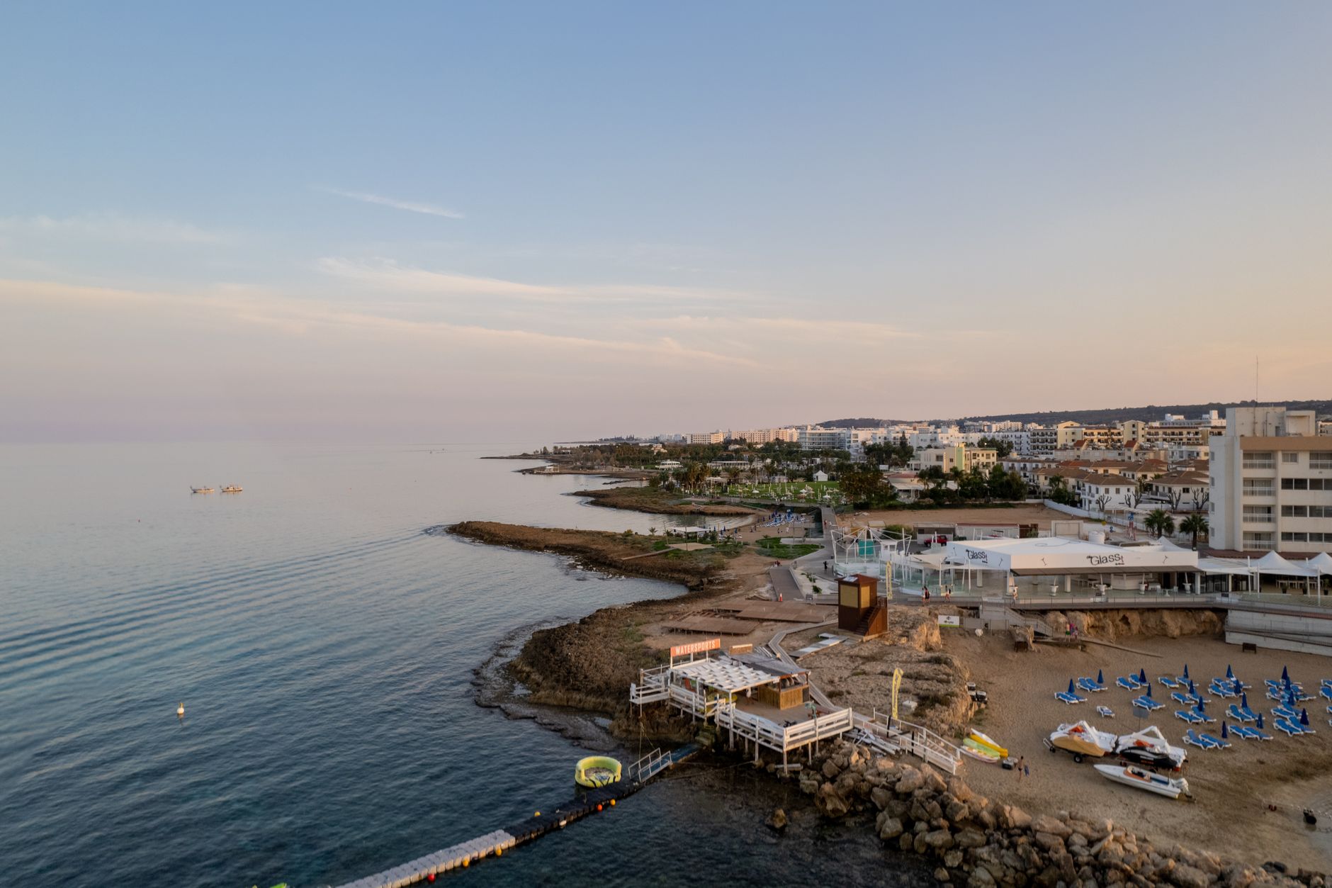 Aerial view of a Cypriot resort coastline at dusk, with a rocky shore, a watersports jetty on stilts, rows of blue sunloungers on the sand and low white apartment blocks running into the distance