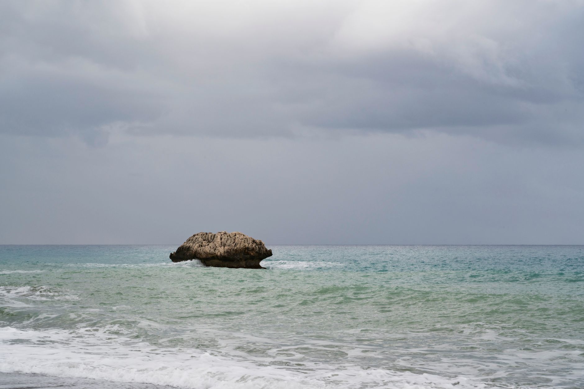 A single weathered limestone rock standing in shallow turquoise sea just off a Cyprus beach, with surf breaking around it under a heavy grey sky