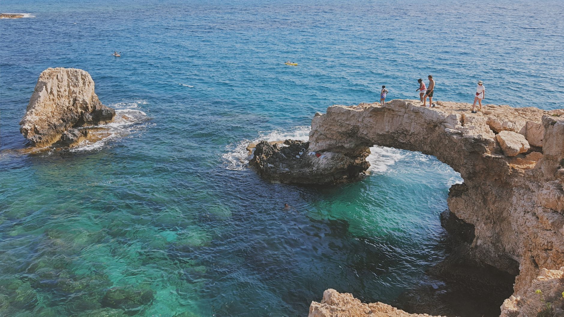 Four people standing on a natural limestone sea arch above clear turquoise water on the rocky Cyprus coast, with kayakers further out