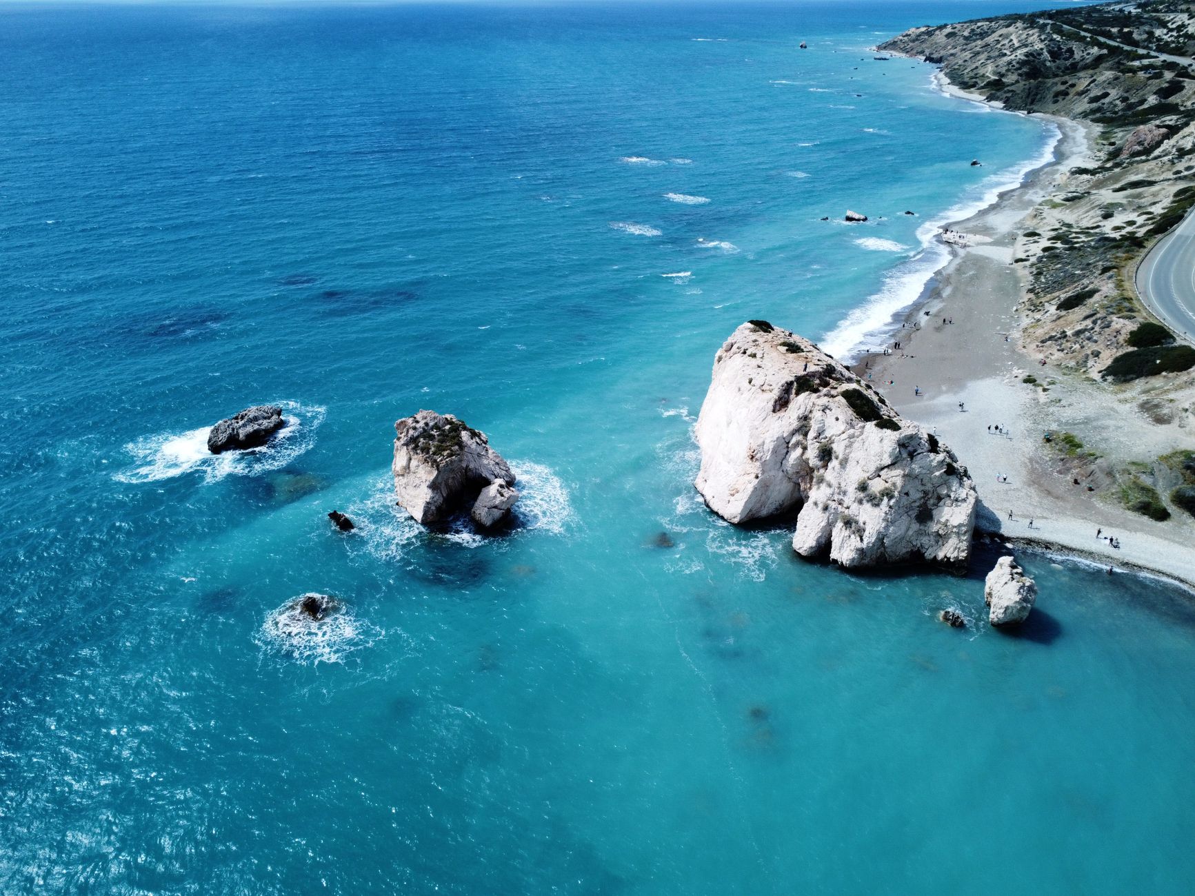 Aerial view of two large white sea stacks in bright turquoise water beside a pebble beach and a coastal road on the south-west coast of Cyprus