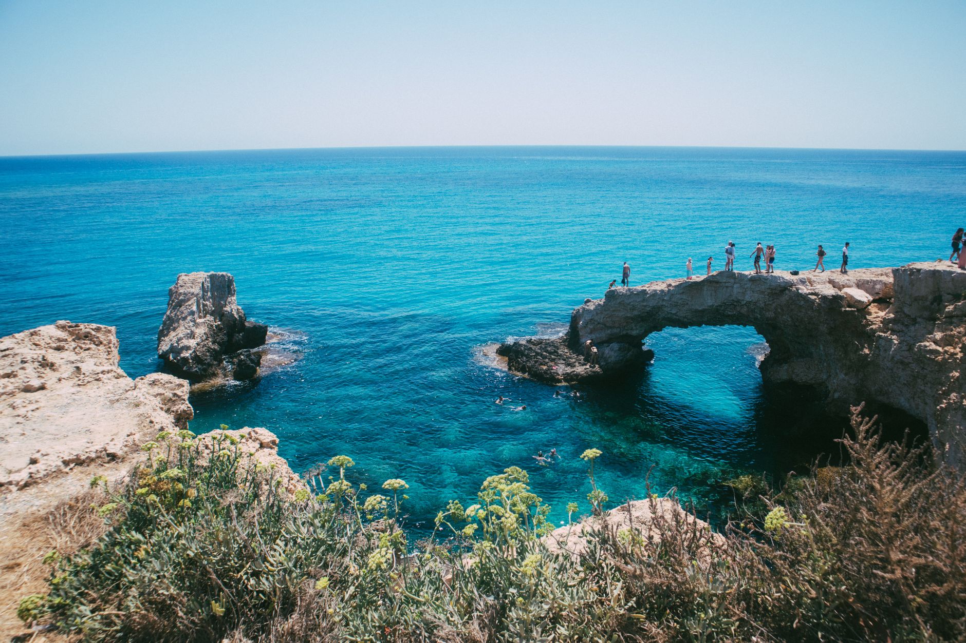 A natural limestone sea arch on the Cyprus coast with people walking across the top of it, the clear turquoise water below full of swimmers