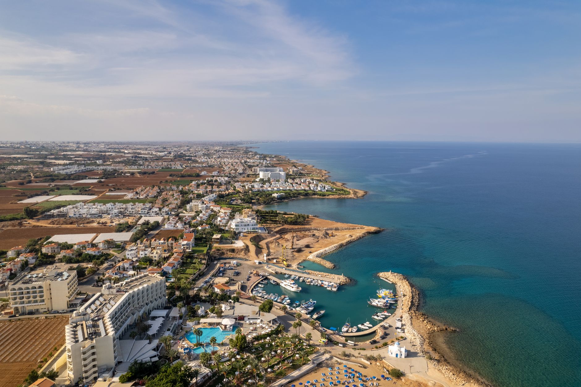 An aerial view of the Cyprus coast, with a small harbour full of fishing boats, a white blue-domed chapel on the headland and rows of blue sun loungers on the sand