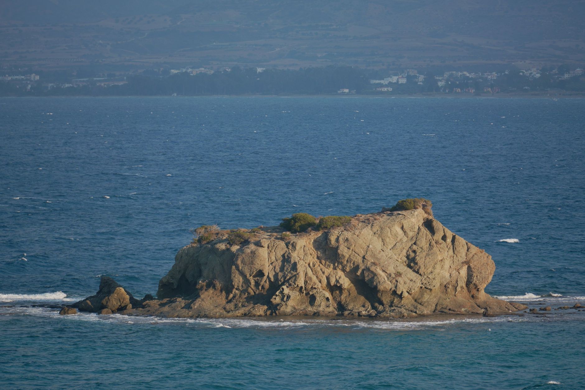 A weathered rock island with scrub on its ridge standing in deep blue open sea, with a hazy coastline of low white buildings and dry hills behind it