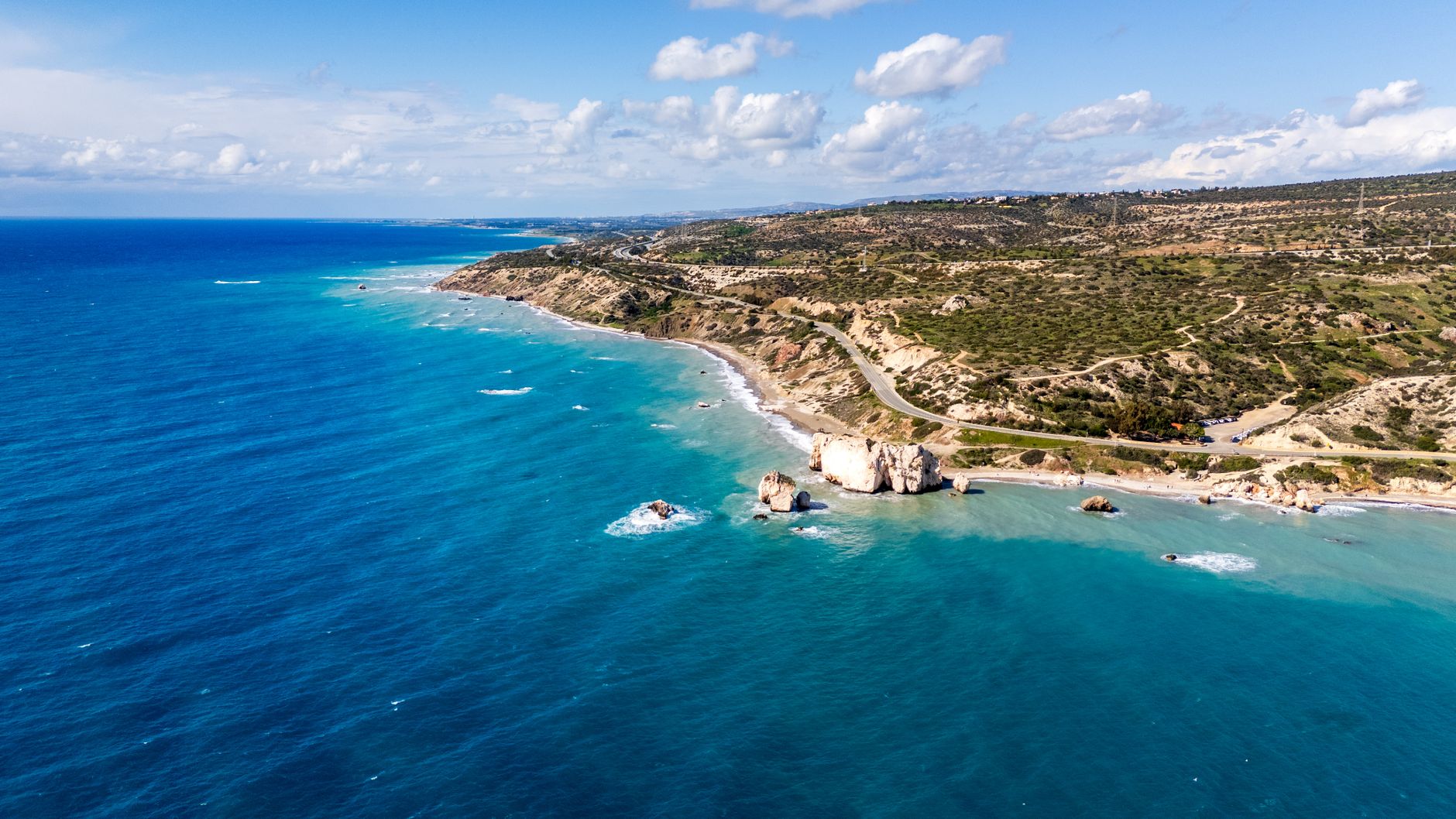Aerial view of the Cyprus south-west coastline, with sea stacks in turquoise shallows, a pale limestone shore and the coast road curving through scrubland above it