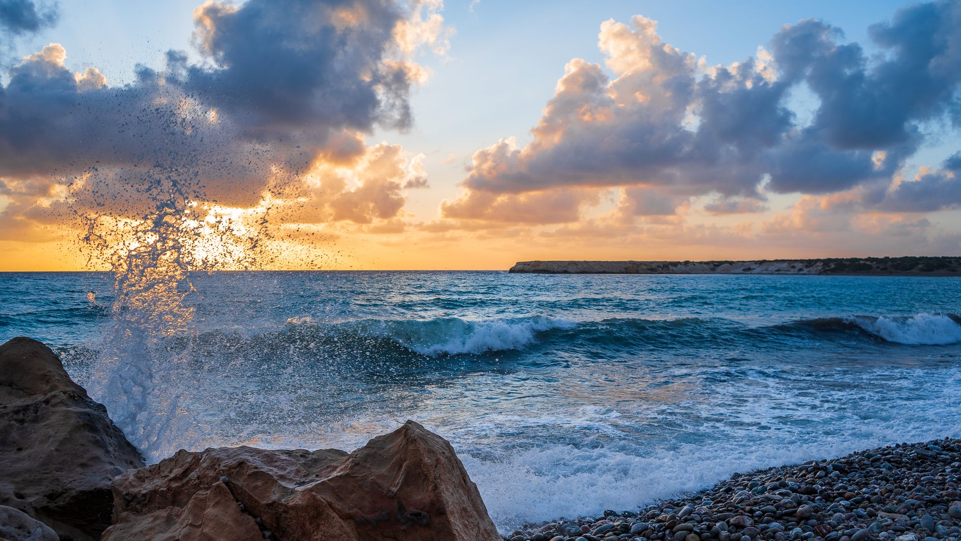 A wave breaking against sandstone rocks on a pebbled Cyprus shore at sunset, with a low headland on the horizon
