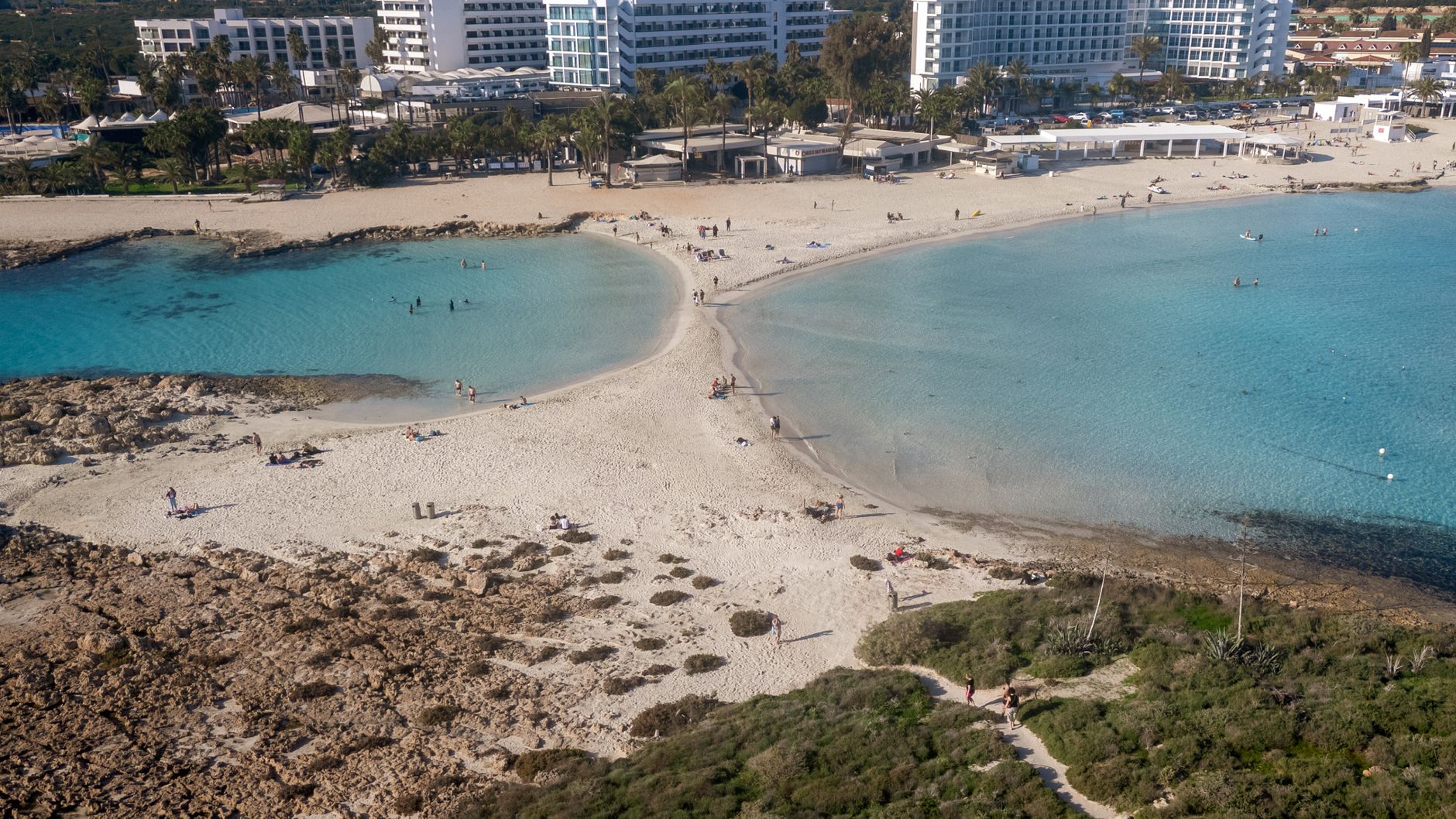 חוף ניסי באיה נאפה ממעוף הציפור: לשון חול בהירה שיוצאת אל אי סלעי בין שני מפרצים של מים טורקיזיים צלולים, עם מלונות ודקלים לאורך החוף