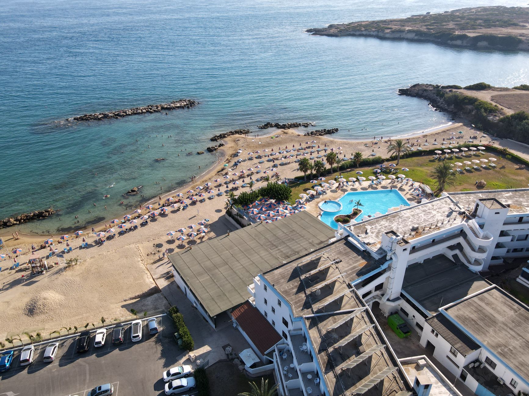 Veduta aerea di una spiaggia cipriota di sabbia chiara con file di ombrelloni e lettini, la piscina di un albergo alle spalle e un promontorio roccioso che entra nell'acqua calma
