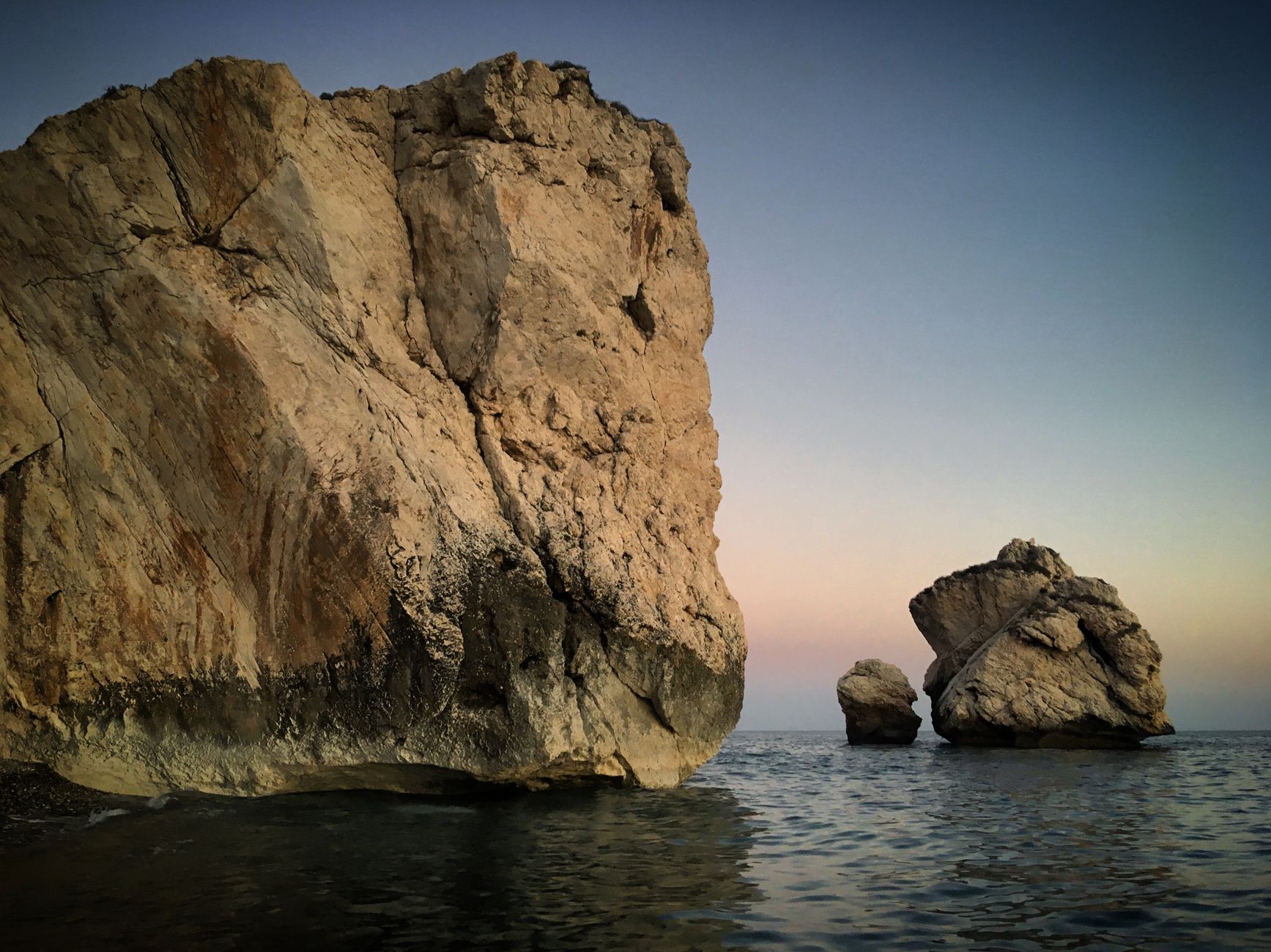 A tall pale limestone rock rising straight out of shallow sea at dusk, with two smaller stacks further out and a pink and blue sky behind them