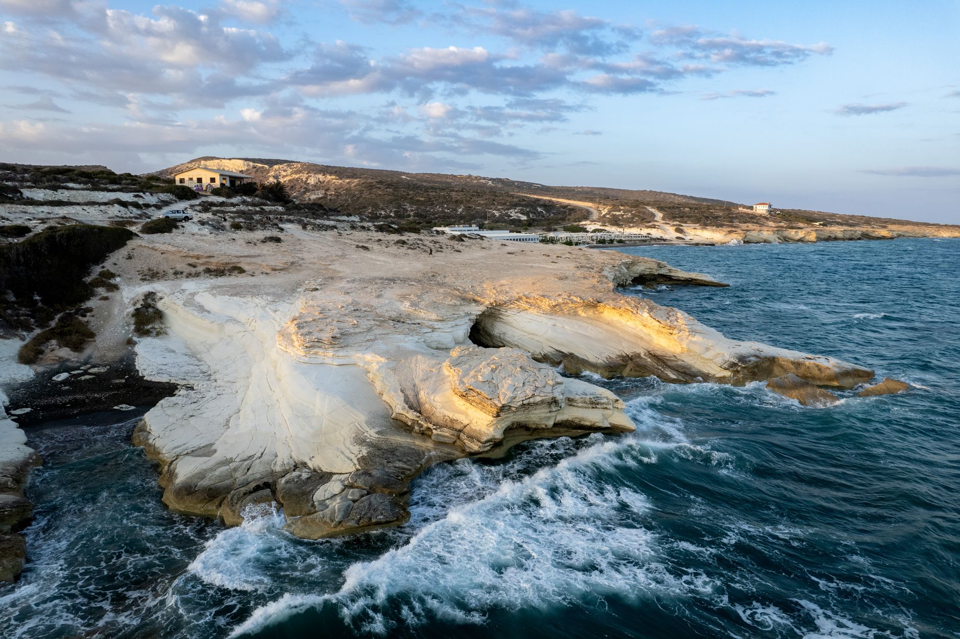 A low aerial view at golden hour of a white chalk headland layered like pastry, waves breaking around its base in the deep blue sea, with a scrubby ridge, a low flat-roofed building and a parked car on the clifftop behind