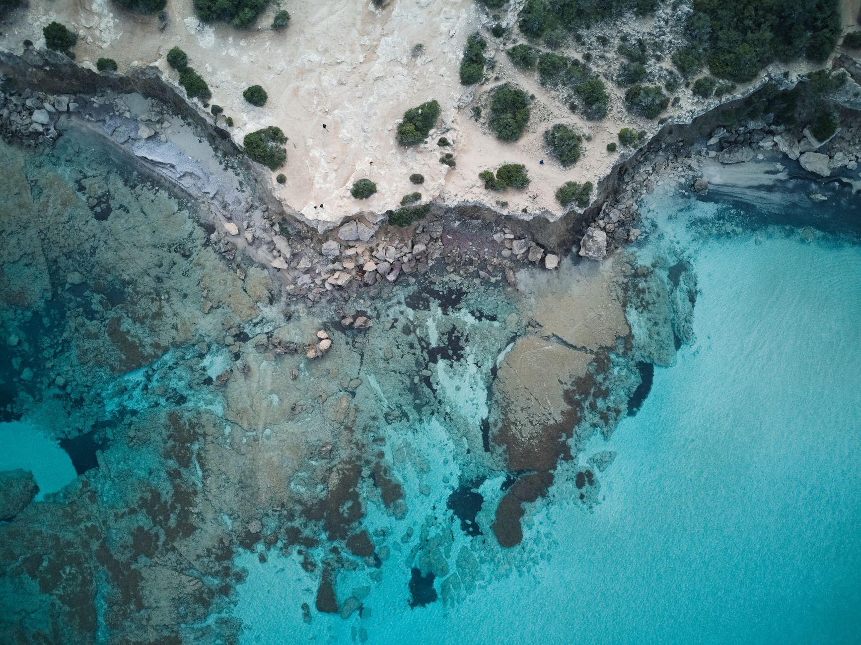 A stretch of Cypriot coast photographed straight down from the air, pale limestone and low green scrub meeting clear turquoise shallows over a rocky seabed