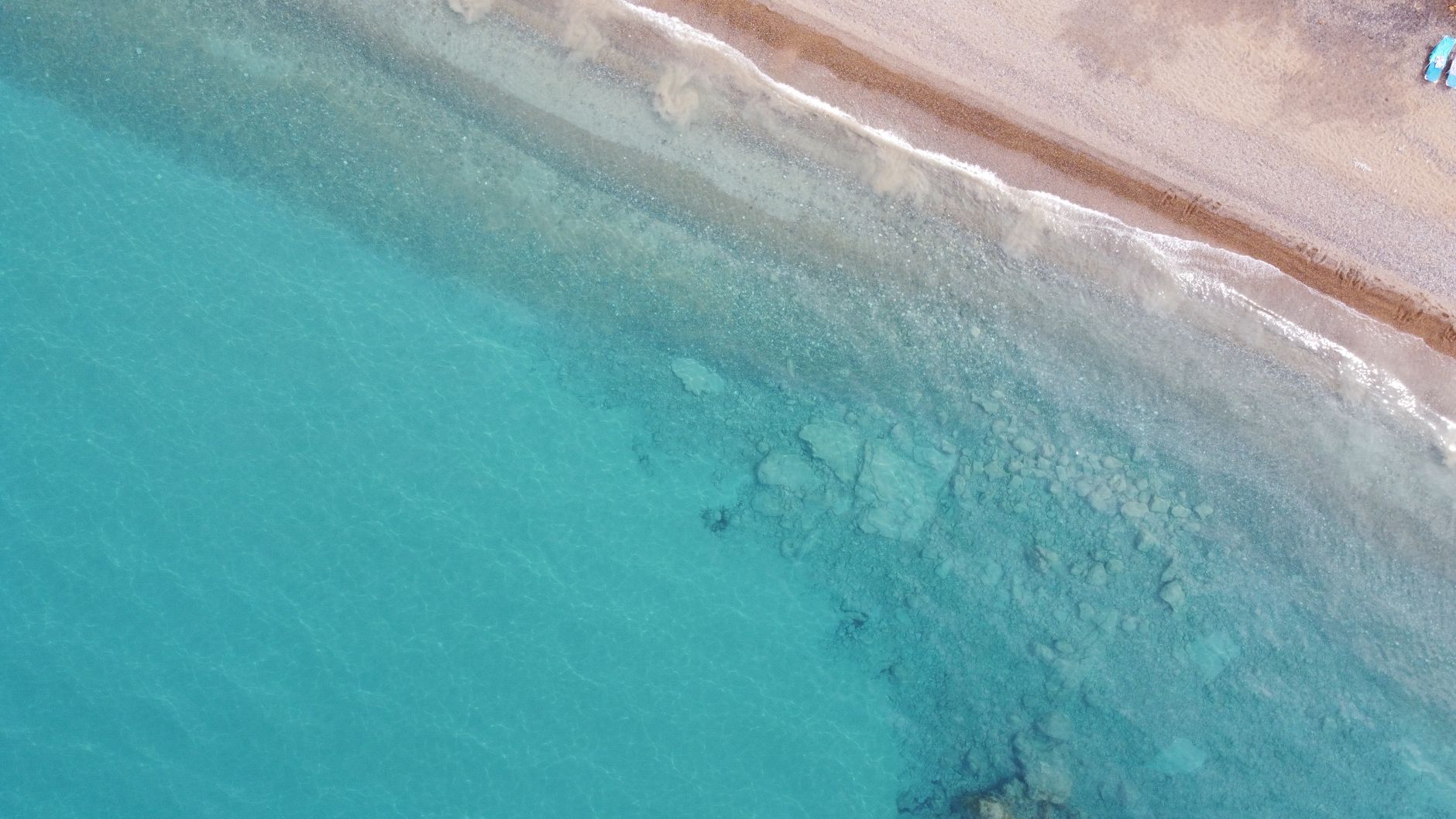 Straight-down aerial view of a Cyprus shoreline: turquoise shallow water over pale submerged rocks meeting a narrow strip of coarse sand, with two small blue boats pulled up at the top edge