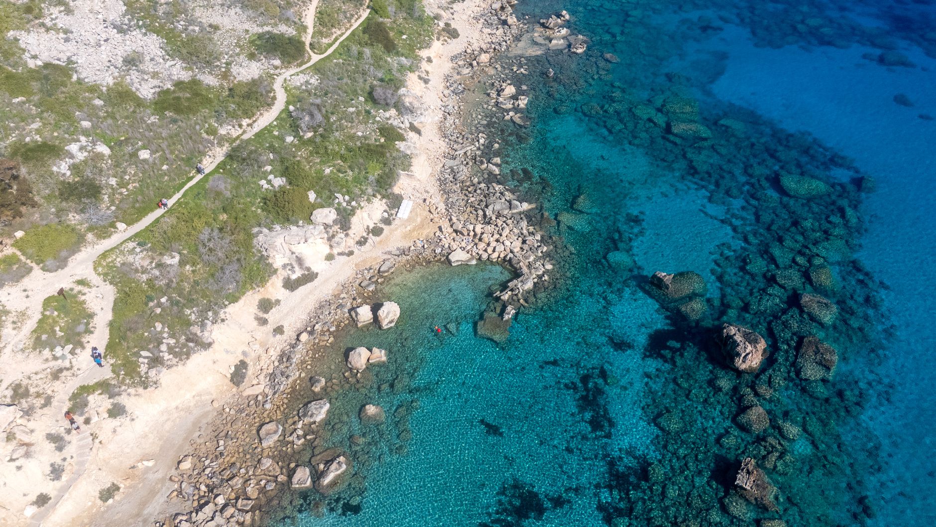 An aerial view of a Cyprus coastline, pale limestone rocks and low scrub meeting turquoise shallows that darken to deep blue, with a footpath running along the headland and two swimmers near the shore