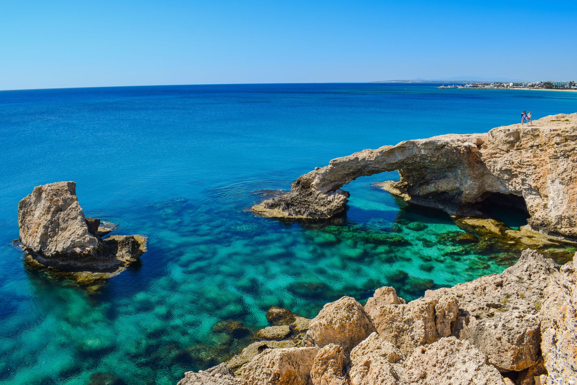 A natural rock arch over turquoise shallows at Cape Greco in Cyprus, with two people standing on the cliff above