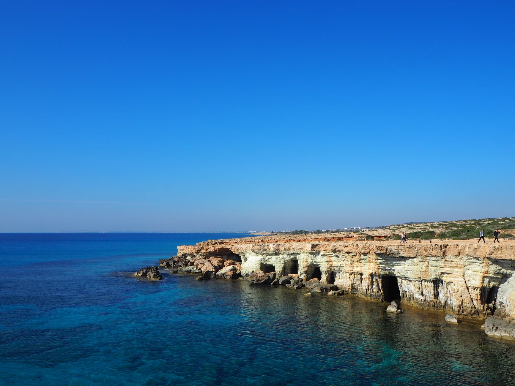 A low limestone cliff riddled with sea caves running out into deep blue Mediterranean water, with two people walking along the clifftop path in the distance