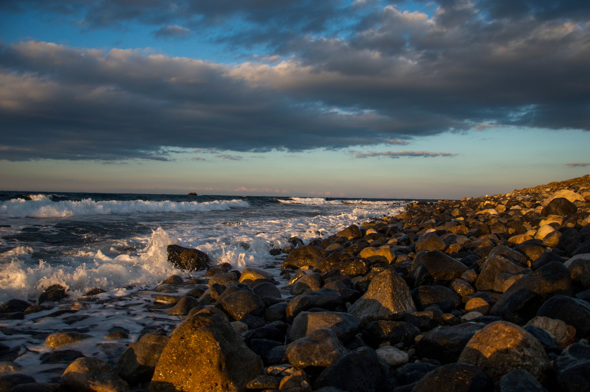 Waves breaking over a bank of large rounded boulders on a Mediterranean shore at sunset, the wet stones lit gold beneath a wide band of grey and blue cloud