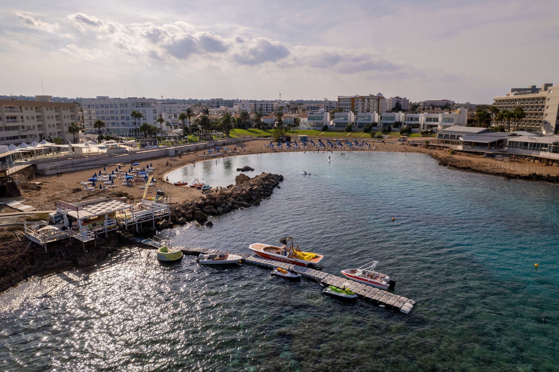 An aerial view of a sheltered sandy bay on the Cyprus coast, with rows of blue sun loungers, a floating pontoon carrying speedboats and jet skis, and low white hotels behind the beach