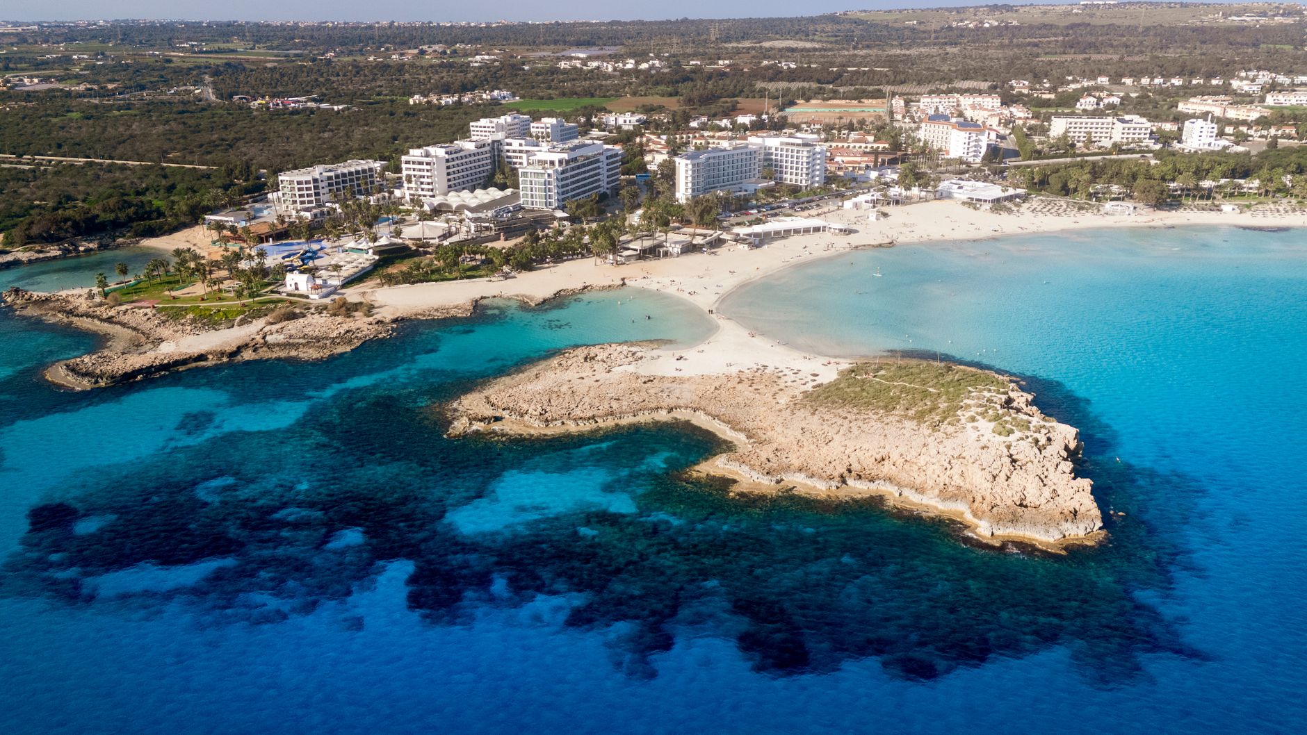 Aerial view of a wide sandy bay in Cyprus with a rocky islet in the turquoise shallows and low-rise white hotels along the shore