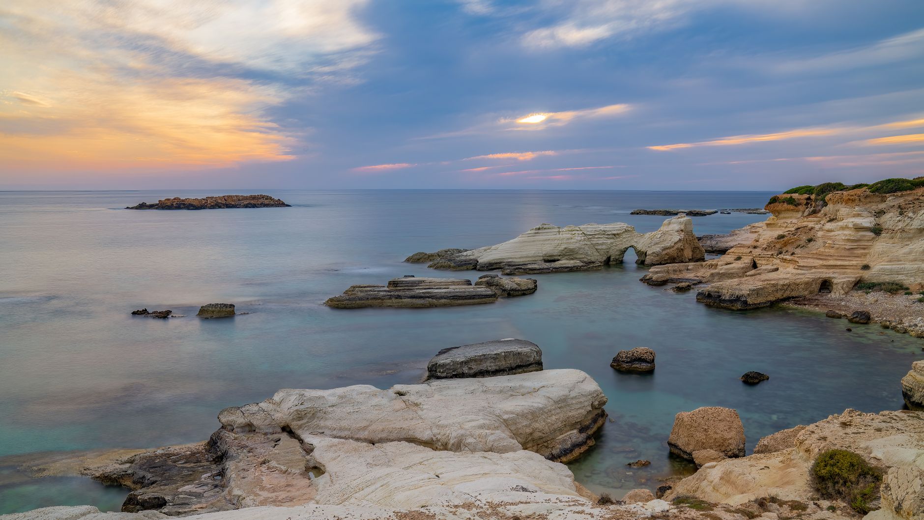 Pale limestone rock formations and a natural arch in shallow turquoise water on the Cyprus coast at dusk, with an orange and violet sky above a flat sea