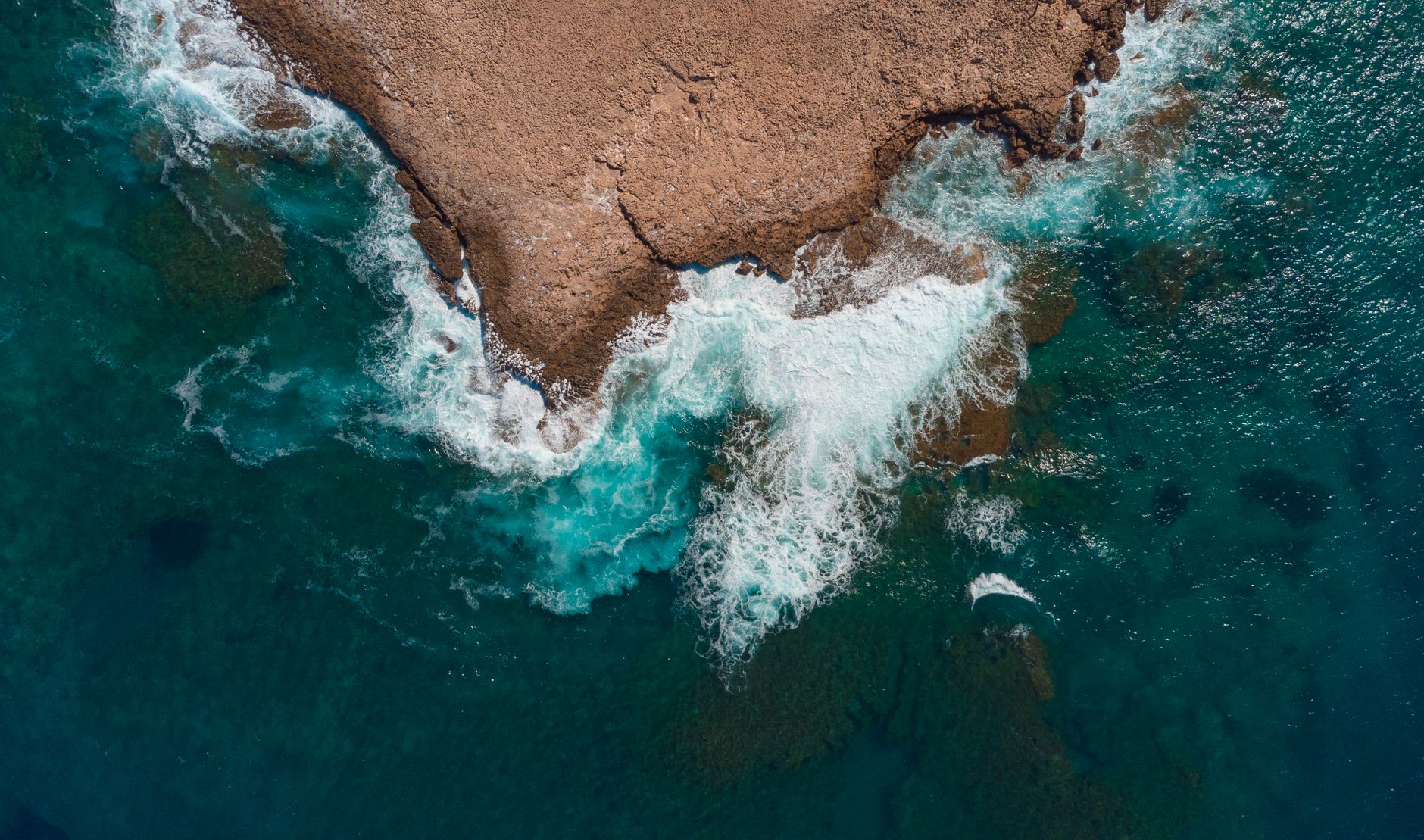 A drone photograph looking straight down at a tan rock headland reaching into deep teal water, with white surf wrapping around its edges and submerged reef showing green beneath the surface