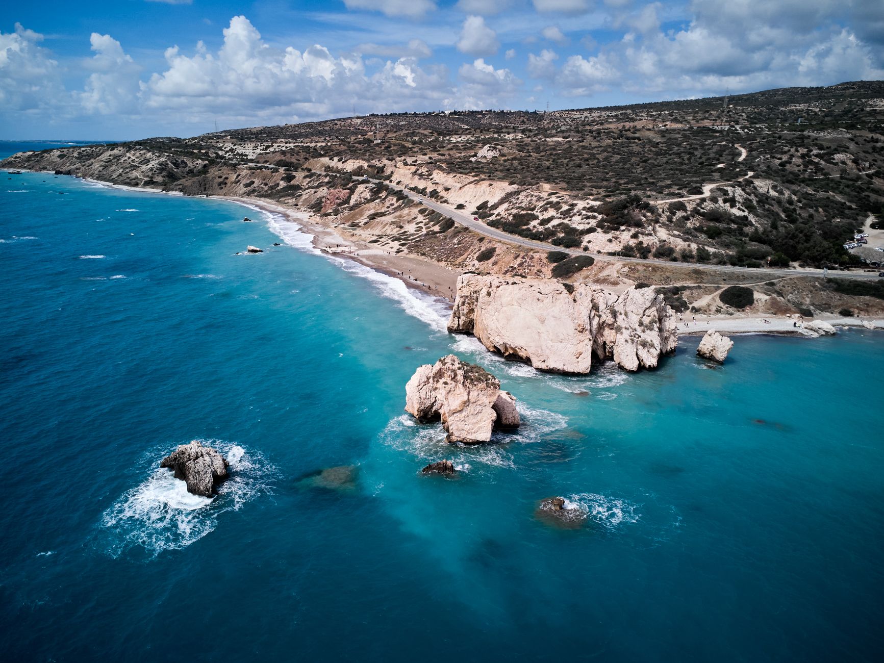Aerial view of a Cyprus coastline where pale limestone sea stacks rise out of turquoise water beside a narrow pebble beach, with a road climbing through dry scrub hills under a cloudy sky