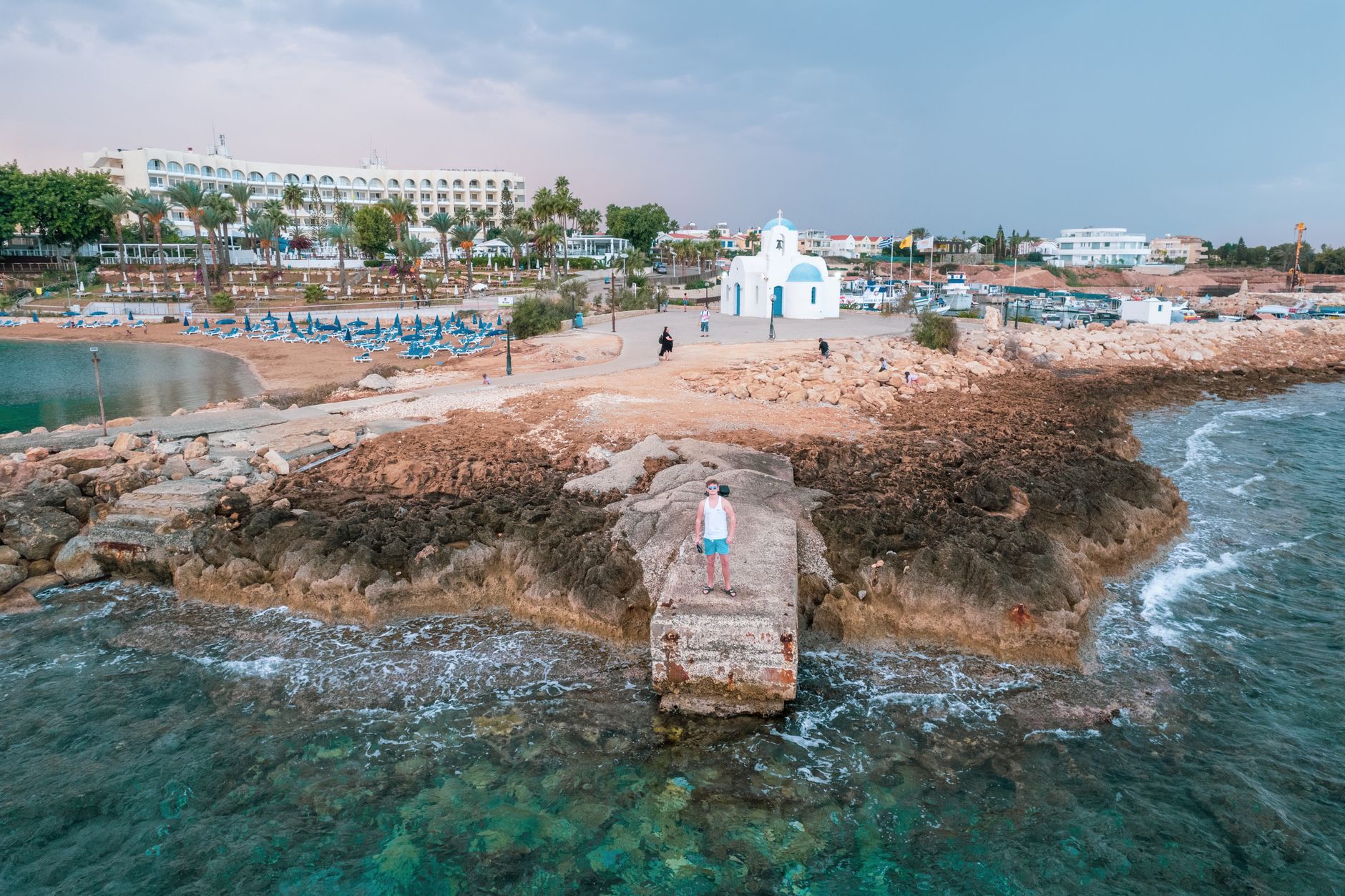 A small white chapel with a blue dome on a rocky headland beside a Cyprus fishing harbour, with a man standing on a concrete jetty above clear turquoise water
