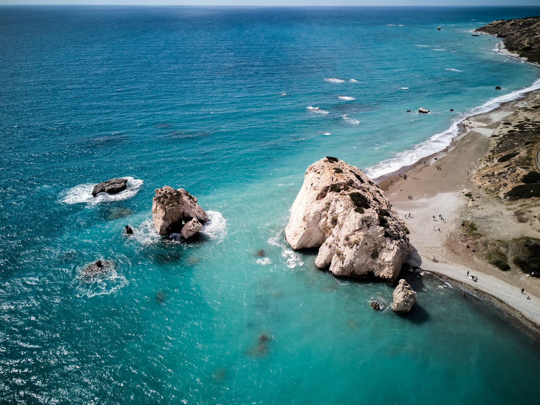 Luftaufnahme heller Kalkfelsen, die aus türkisfarbenem Wasser neben einem geschwungenen Kiesstrand aufragen, dahinter trockene Buschhügel