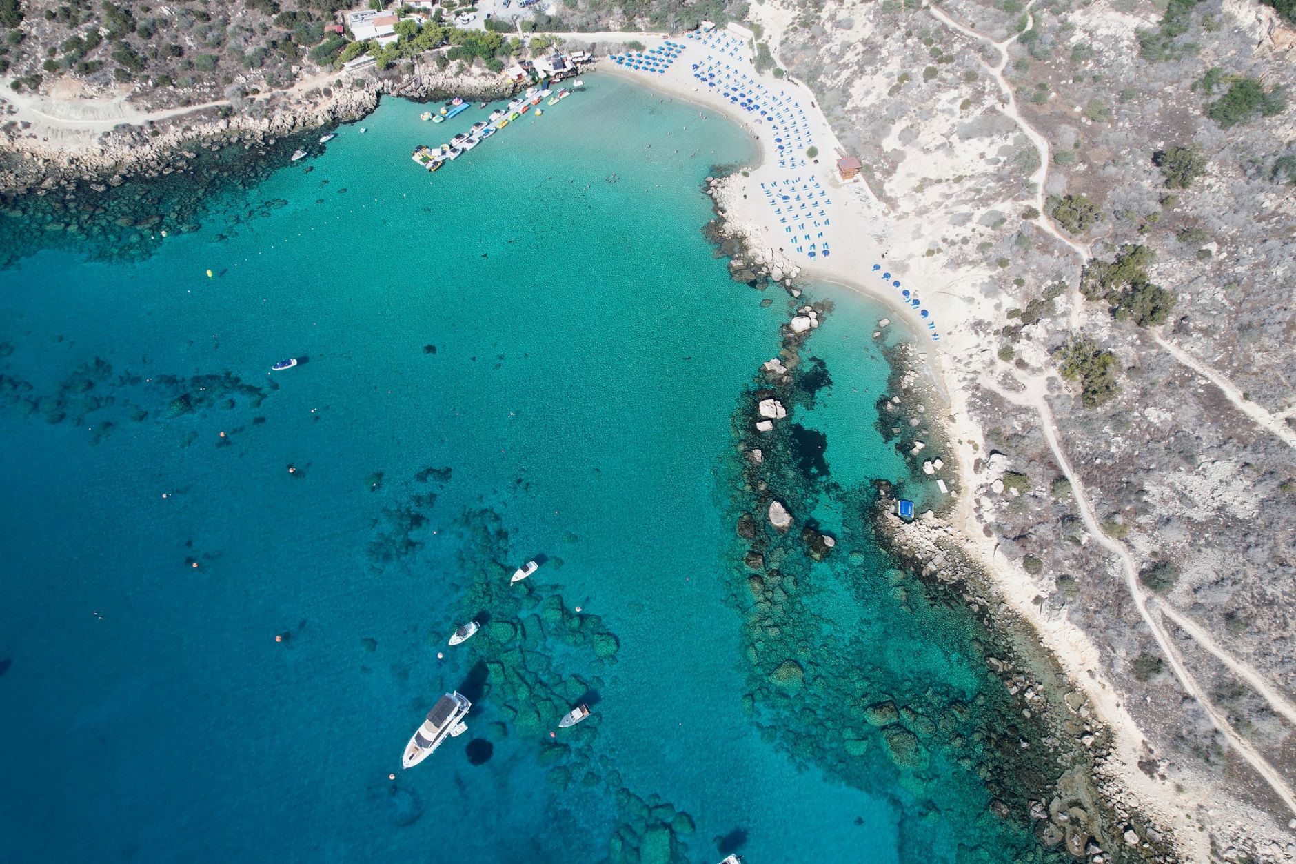 Aerial view of a turquoise Cyprus bay with rows of blue sunbeds on a small sandy beach, moored boats and a dry rocky headland