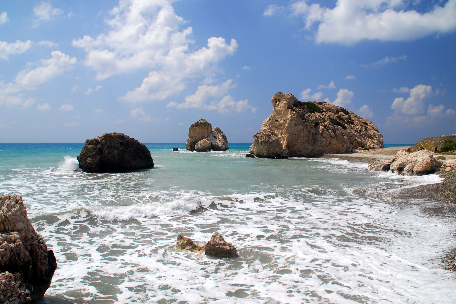 Waves breaking over pale limestone sea stacks in clear turquoise water at Petra tou Romiou on the south-west coast of Cyprus, under a blue sky with scattered cloud