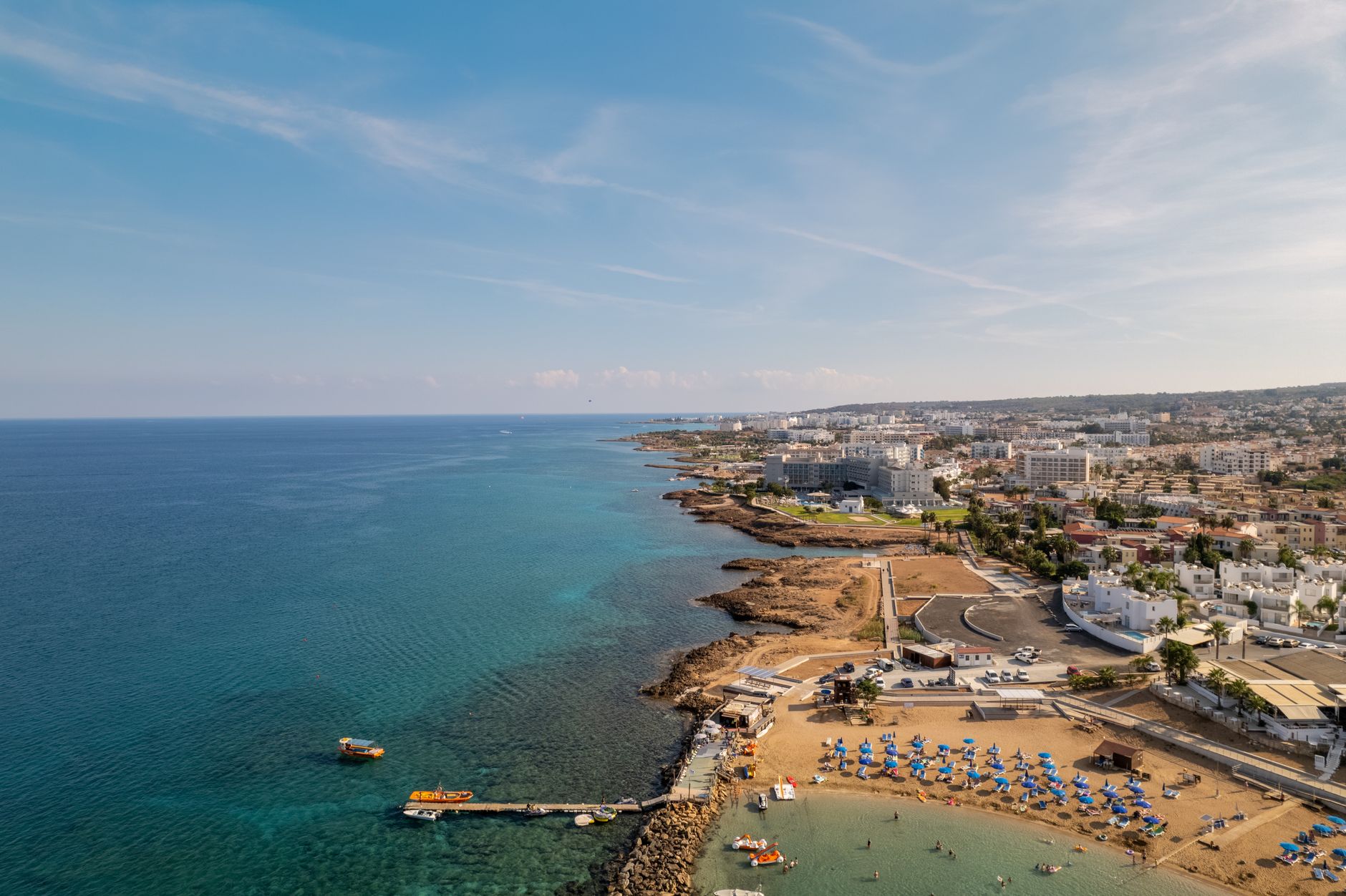 A sandy Cyprus resort bay with rows of blue parasols, a jetty and small boats, and low white hotels running along the coast behind