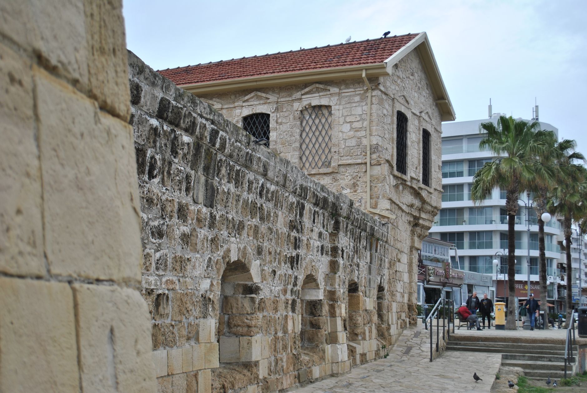 A weathered limestone fort wall in Cyprus with lattice-barred windows under a tiled roof, a cobbled path running along its foot, and palm trees and a curved white modern building behind it