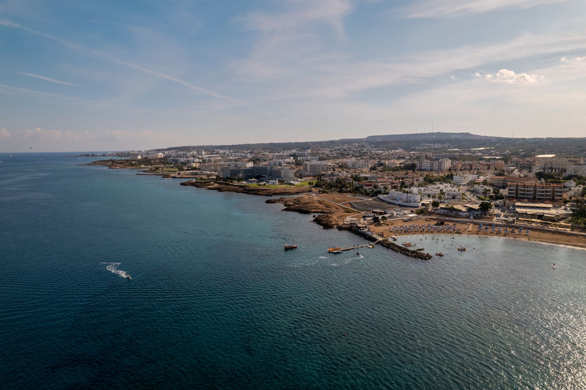 The Cyprus coastline from the air, a low white resort town along a headland with a busy beach and a jetty reaching into deep blue water