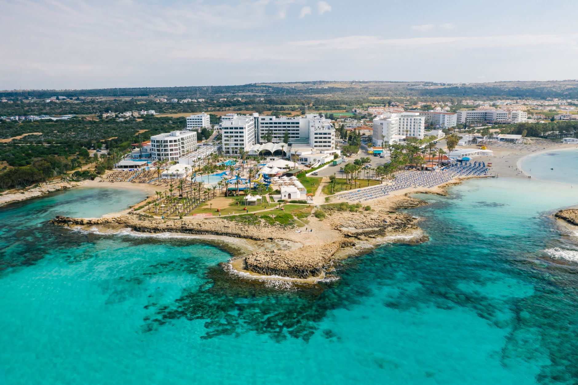 A Cyprus coastal resort strip photographed from the air: turquoise shallows over rock, a low headland of pale stone, palm-lined lawns and rows of blue sunbeds in front of white low-rise hotels