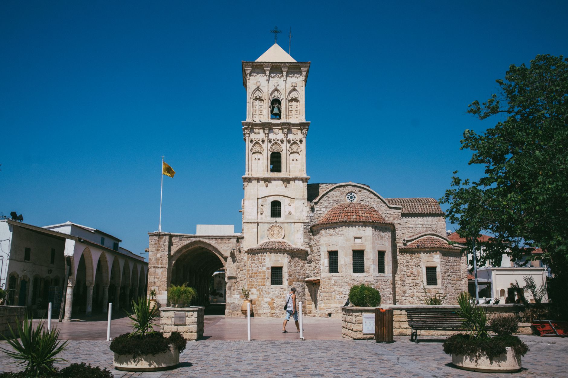 A honey-coloured stone church with a tall carved bell tower on a paved Cypriot town square, under a cloudless deep-blue sky