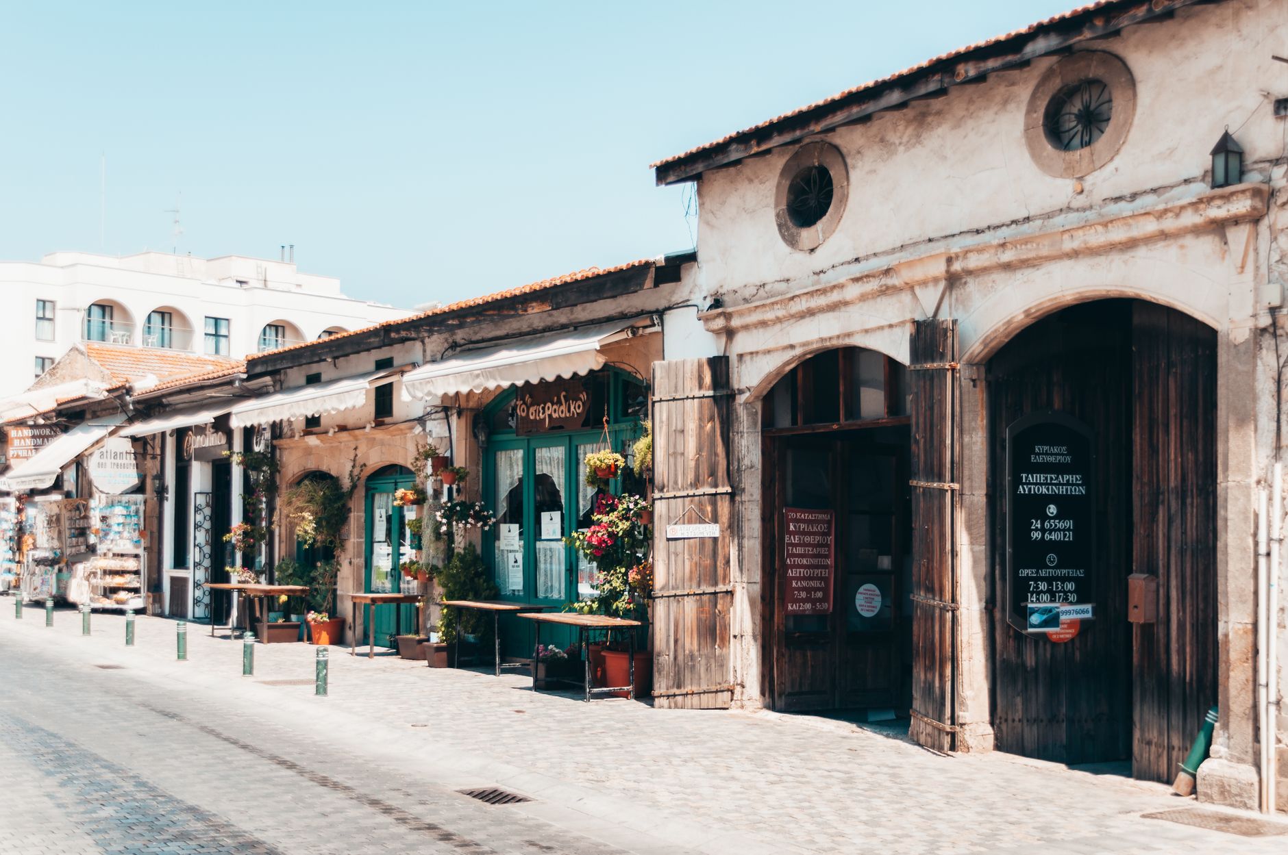 A sunlit old-town street in Cyprus lined with low stone and stucco shopfronts, terracotta roof tiles, canvas awnings and Greek-lettered signboards beside two tall wooden doors