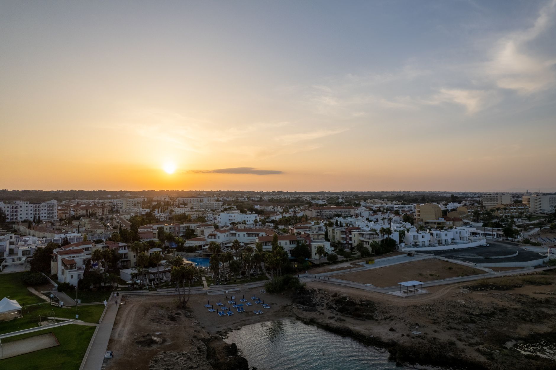 A Cyprus coastal town at sunset seen from the air, white apartment blocks and palm gardens running down to a small sandy cove
