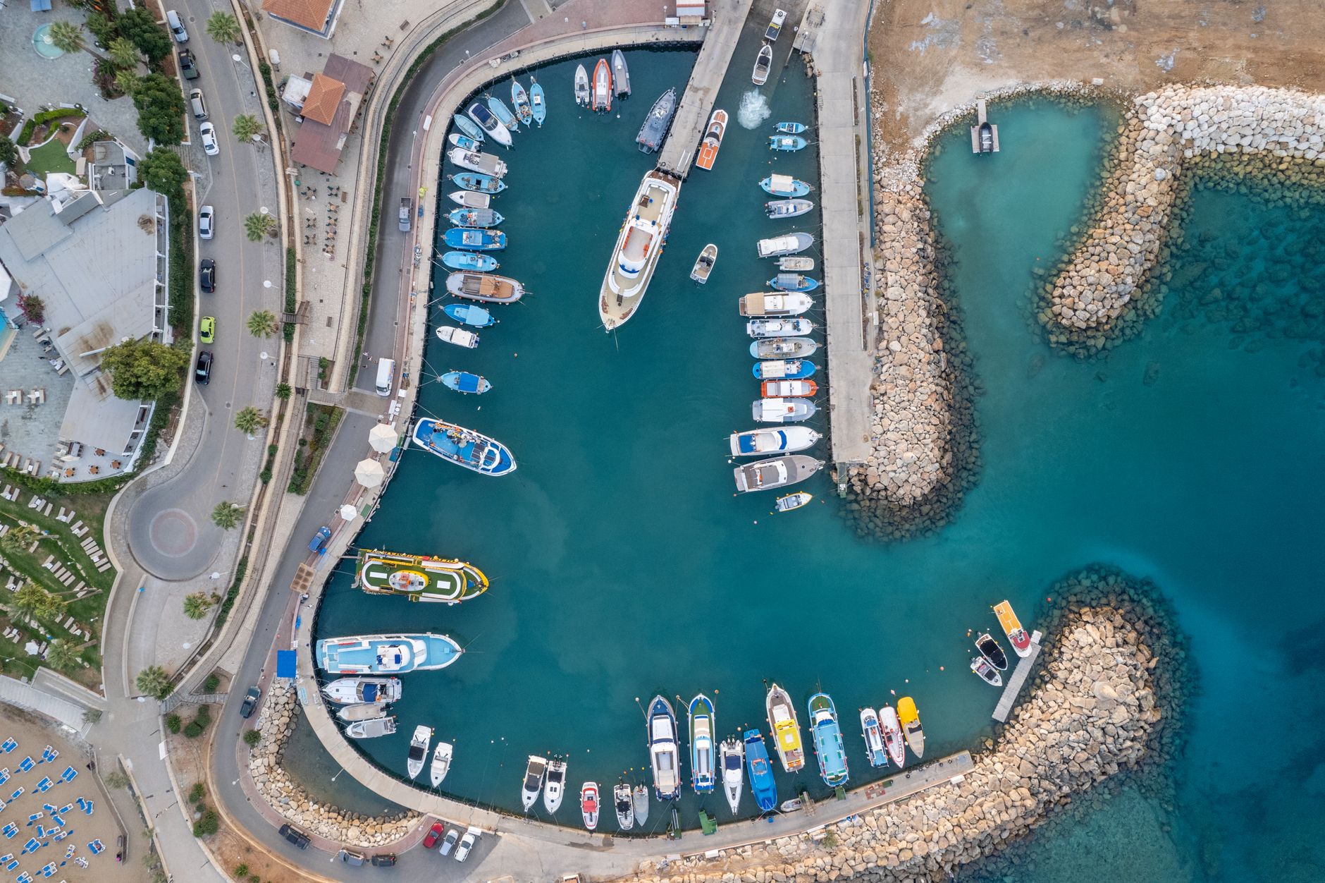 Aerial view looking straight down on a small Mediterranean marina in Cyprus, packed with fishing boats and motor cruisers between a rubble breakwater and a coastal road