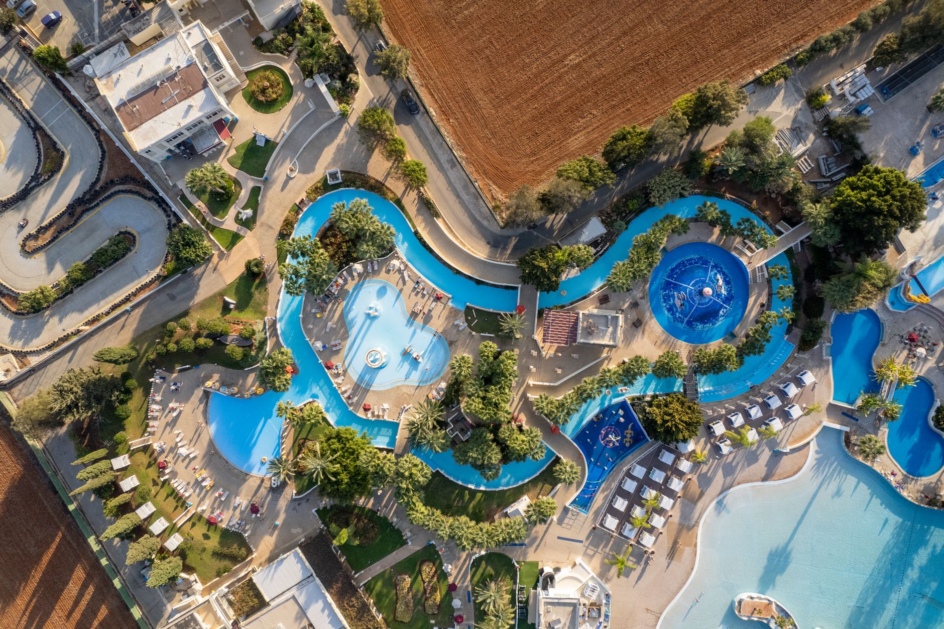 Overhead aerial view of a Mediterranean resort complex: a winding blue lazy river and several free-form swimming pools threading between palm trees, sun loungers and white cabanas