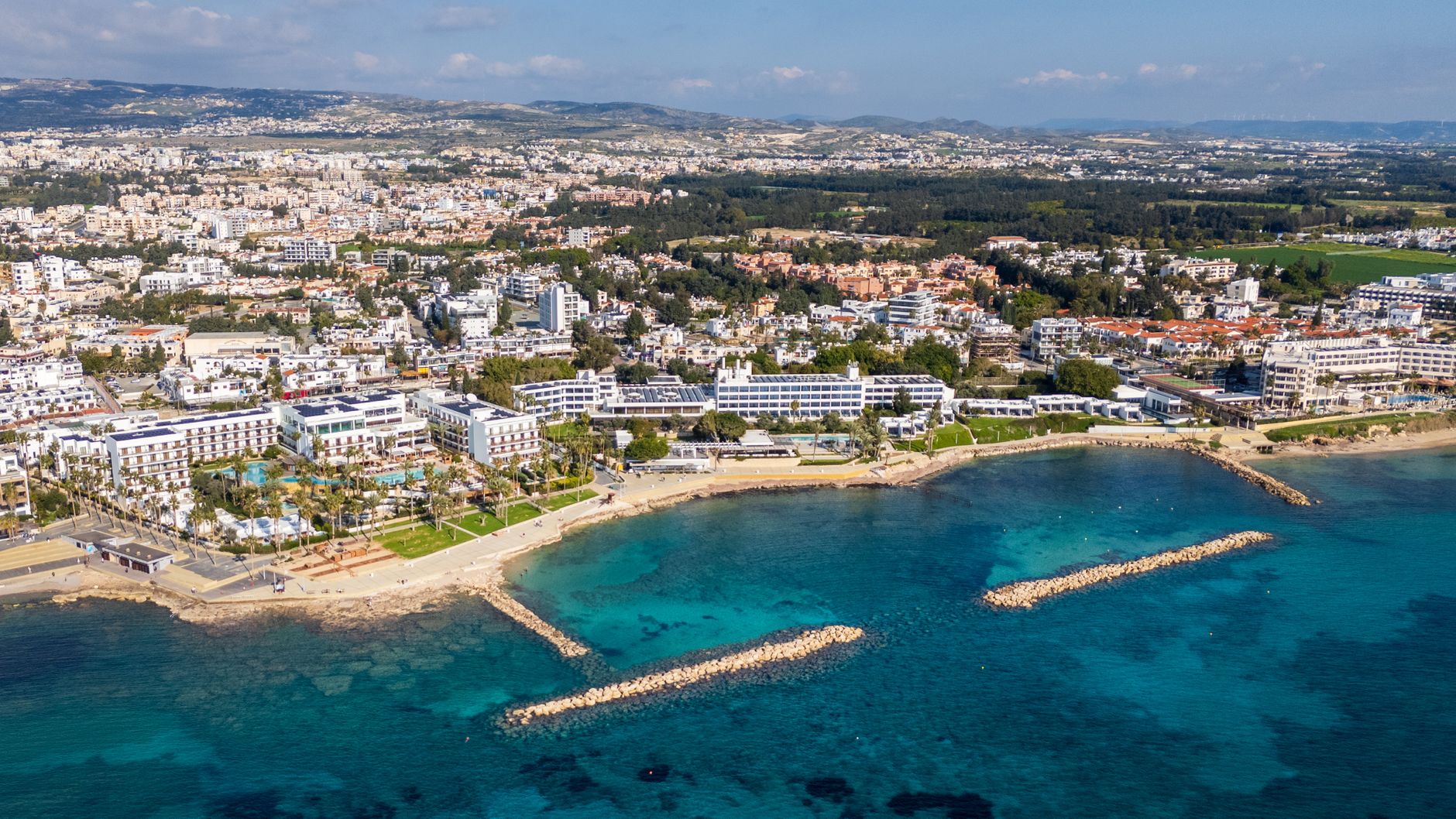 A Cyprus coastal city seen from the air, with low white hotels along a palm-lined seafront and stone breakwaters in clear turquoise water