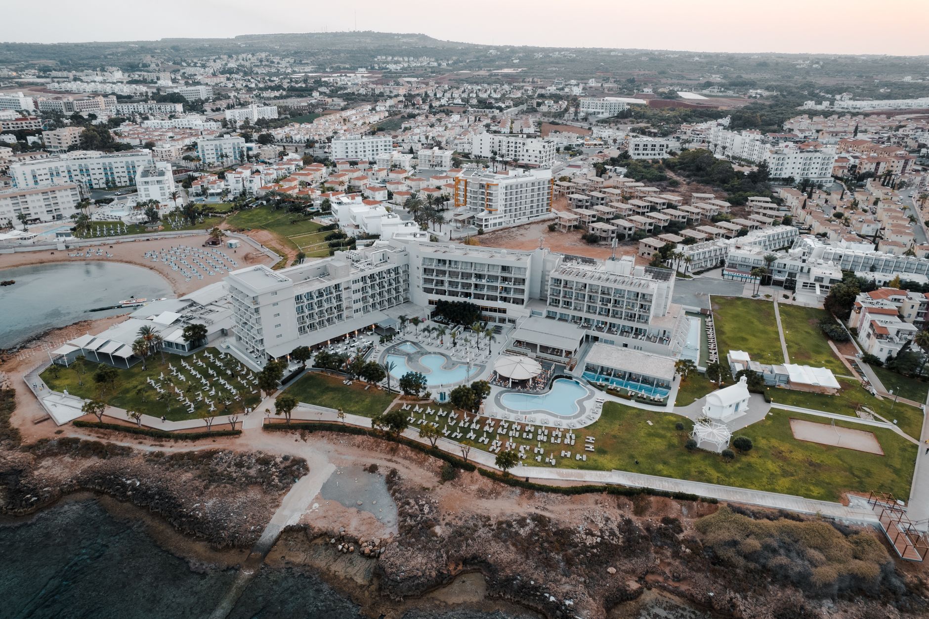 Aerial view of a Cyprus coastal resort district at dusk, with a low white hotel and pools on the shoreline and villas rising behind it