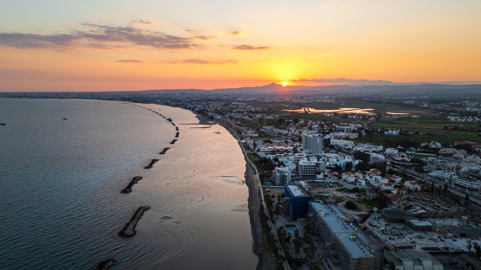Eine zyprische Küstenstadt aus der Luft im Sonnenuntergang, das Meer auf der einen Seite, niedrige weiße Häuser landeinwärts und Berge am Horizont